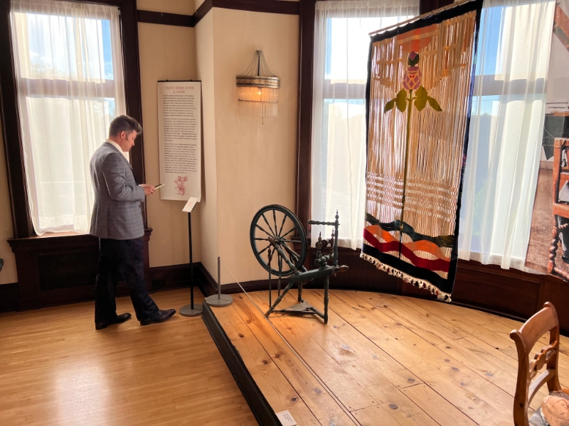 A visitor reads exhibit content on a smartphone beside a historic spinning wheel and textile